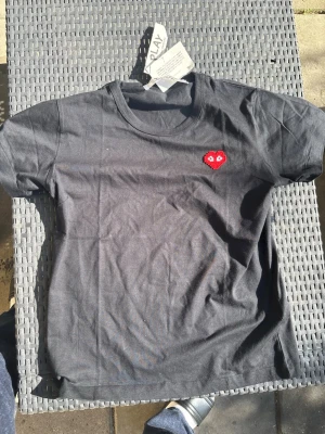 Svart t-shirt med rött hjärta - Snygg svart t-shirt med ett litet rött hjärta med ögon broderat på bröstet. Klassisk rund halsringning och korta ärmar. Perfekt till jeans eller shorts för en enkel men cool look.