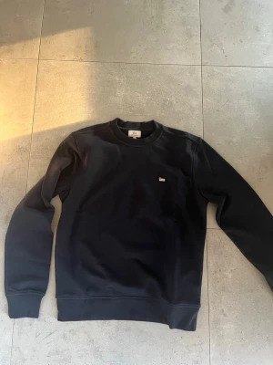 Woolrich sweatshirt oanvänd - En stilren oanvänd mörkblå sweatshirt från Woolrich i toppskick med rund halsringning och diskret logga på bröstet. Tröjan har ribbade muddar vid ärmslut och nederkant. Perfekt för en avslappnad och snygg look.