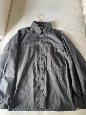Grå oversized overshirt med fickor - Snygg grå overshirt i oversized modell med stora bröstfickor och knäppning framtill. Skjortan har klassisk krage och långa ärmar med knapp vid ärmslut. Perfekt att bära som lager över t-shirt. Tillverkad i bomull.