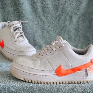 Vita Nike sneakers med orange swoosh - Säljer ett par vita Nike sneakers med en iögonfallande orange swoosh på sidan. Skorna har klassisk låg profil, perforerad tåbox och vita snören. Yttermaterialet är i läder och sulan är vit med diskret mönster. Perfekta för dig som gillar streetwear och vill sticka ut.