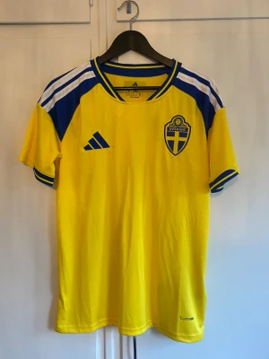 Svensk landslagströja Adidas gul/blå - Gul svensk landslagströja från Adidas med blå och vita detaljer på axlar och ärmslut. Broderad Sverige-logga på bröstet och Adidas-logga. Tillverkad i lätt och ventilerande material, perfekt för fotboll eller träning.