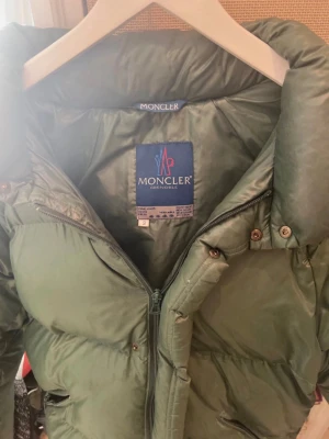 Grön dunjacka från Moncler - Snygg grön dunjacka från Moncler Grenoble med hög krage och dragkedja framtill. Jackan har klassisk Moncler-logga på ärmen och är tillverkad i glansigt material som ger en exklusiv känsla. Perfekt för kalla vinterdagar.