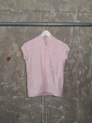 Ljusrosa väst från Fred Perry - Ljusrosa väst från Fred Perry i mjuk frotté med dragkedja framtill och två fickor. Västen har korta ärmar, ribbad krage och ett broderat Fred Perry-logotyp på bröstet. 