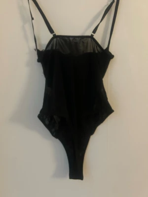 Svart mesh body med spetsdetaljer - Snygg svart body i mesh med tunna axelband och spetsdetaljer på sidorna. Materialet är genomskinligt upptill och ger en elegant look. Perfekt för dig som vill ha något stilrent och trendigt i underklädesgarderoben.