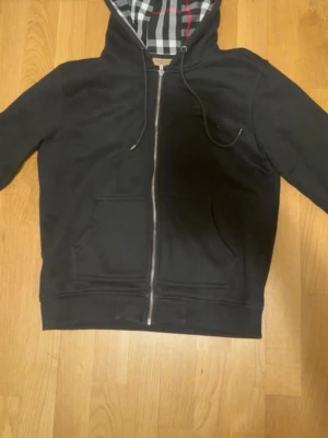 Svart hoodie med rutigt foder - Säljer en svart hoodie med dragkedja och känguruficka. Huvan har ett snyggt rutigt foder i svart, vitt och rött. Klassisk passform och dragsko i huvan. Perfekt för dig som gillar stilrena detaljer och vill ha något bekvämt till vardags.