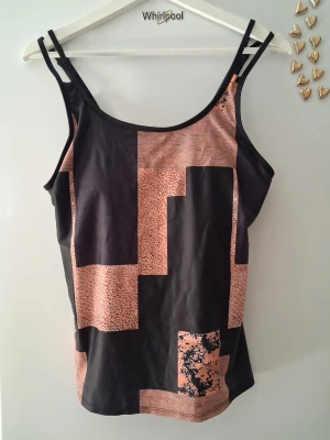 Svart och rosa/brun tankini med mönster - Säljer en snygg tankini i svart med rosa geometriskt mönster. Modellen har dubbla smala axelband och justerbara band baktill för bästa passform. Materialet är stretchigt och snabbtorkande, perfekt för stranden eller poolen.