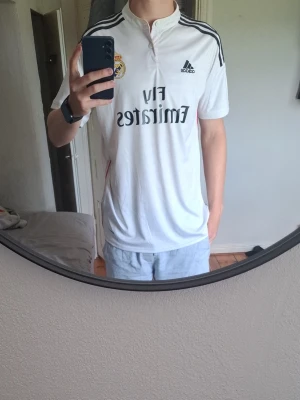 Vit Real Madrid RAMOS - Säljer en vit Real Madrid matchtröja från Adidas med klubbmärke och svarta detaljer på axlarna. Tröjan har korta ärmar, ståkrage med knapp och tryckt sponsorlogga på bröstet. Perfekt för dig som älskar fotboll och vill representera ditt lag.