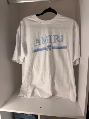 Vit AMIRI t-shirt med blått tryck - Snygg vit t-shirt från AMIRI med ljusblått tryck på bröstet och klassisk rund hals. T-shirten har korta ärmar och är gjord i mjuk bomull, perfekt för en clean och stilren look.