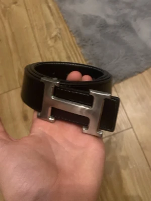 Svart bälte med silverfärgat H-spänne - Stilrent svart bälte med ett stort, silverfärgat H-format spänne. Bältet har vita sömmar längs kanterna och är tillverkat i skinn. Perfekt accessoar för att lyfta din outfit med en klassisk touch.