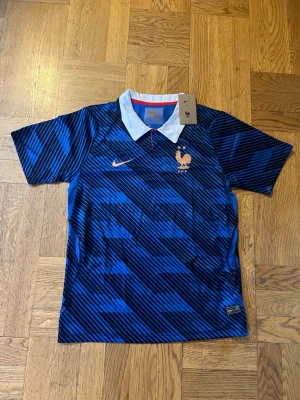 Blå Nike fotbollströja Frankrike - Snygg blå fotbollströja från Nike med Frankrikes landslagslogga på bröstet. Tröjan har ett mönster av svarta diagonala ränder, vit krage med röd detalj och korta ärmar. Perfekt för dig som älskar fotboll och vill visa ditt stöd för Frankrike.