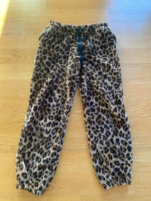Leopardmönstrade mjuka loungewearbyxor - Supermysiga loungewearbyxor med leopardmönster i brunt, svart och beige. Byxorna har resår i midjan och svarta knytband samt resår vid bensluten. Perfekta för chill hemma och riktigt sköna tack vare det fluffiga materialet.