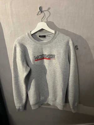 Grå sweatshirt från McKenzie - Snygg grå sweatshirt från McKenzie med tryckt logga i svart, vitt och rött på bröstet. Tröjan har rund halsringning, ribbade muddar vid ärmslut och nederkant samt en klassisk relaxed fit. Perfekt för en casual och sportig stil.