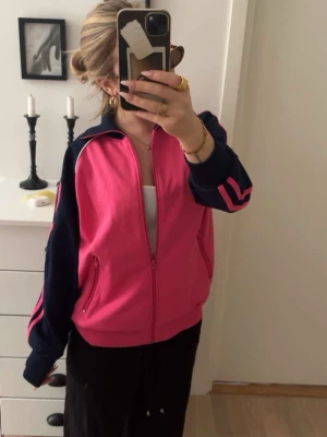 Rosa och marinblå ziptröja - Sportig ziptröja i stark rosa med marinblå ärmar och detaljer. Tröjan har dragkedja framtill, två fickor och rosa ränder längs ärmarna. Perfekt för en avslappnad streetstyle-look. Materialet ser ut att vara bomull eller polyesterblandning.