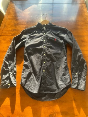 Mörk blå Ralph lauren skjorta - Klassisk mörkblå skjorta med långa ärmar och vita knappar. Skjortan har button-down krage och en liten röd broderad logga på bröstet. Perfekt till jeans eller chinos för en clean look.