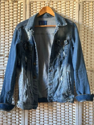 Distressed blå jeansjacka från GAVANI - Snygg blå jeansjacka från GANT med slitna detaljer och klassisk krage. Jackan har knäppning framtill, två bröstfickor med lock och två sidofickor. Perfekt för en avslappnad och trendig look.