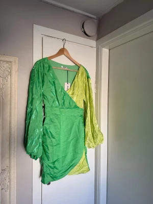 Grön omlottklänning Margaux Dietz x NA-KD - Säljer en kort omlottklänning i två gröna nyanser från Margaux Dietz x NA-KD. Klänningen har ballongärmar, v-ringning och en snygg draperad effekt. Materialet är glansigt och känns lyxigt. Pris kan diskuteras.