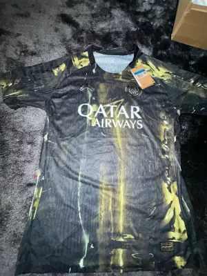 PSG matchtröja svart/gul - Snygg PSG matchtröja i svart med gula och gröna detaljer. Tröjan har korta ärmar, rund hals och Qatar Airways-logga på bröstet. Tillverkad i lätt och ventilerande material, perfekt för fotboll eller träning.