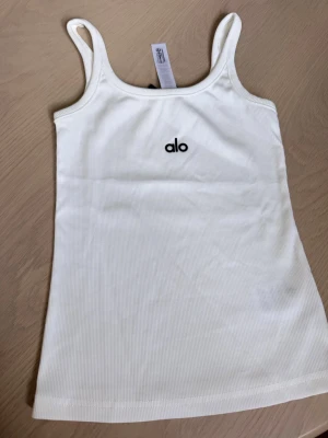 Vit ribbad linnetopp  - Säljer en vit ribbad linnetopp fr med smala axelband och broderad logga framtill. Toppen har en enkel och stilren design som passar perfekt till jeans eller kjol. Materialet är mjukt och stretchigt för en bekväm passform.