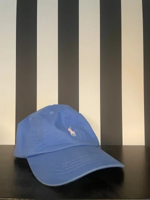 Blå Beach keps från Polo Ralph Lauren - Snygg blå keps från Polo Ralph Lauren med klassisk böjd skärm och justerbart spänne bak. Framtill finns den ikoniska broderade polospelaren i vitt och rosa, och baktill står det 'POLO' broderat i rosa. Tillverkad i bomull för en bekväm känsla.