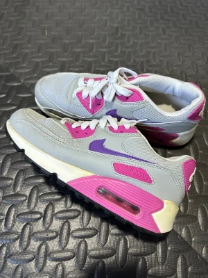 Nike Air Max sneakers grå rosa lila - Nike Air Max sneakers i grått med detaljer i rosa och lila. Klassisk modell med vit sula och synlig Air-enhet. Ovandel i mesh och syntet, snörning framtill och tydlig Nike-logga på sidan och hälen.