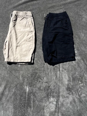 Beige och mörkblå shorts med snörning - Snygga beige och mörkblåa shorts med avslappnad passform och elastisk midja med snörning. Shortsen har två sidofickor och är tillverkade i ett mjukt material som känns skönt mot huden. Perfekta för varma dagar och enkel att matcha med olika stilar. Man får med båda shortsen dom mörkblåa är från Lindex och de beige är från Kappahl Vid frågor hör av er och priset kan alltid diskuteras 