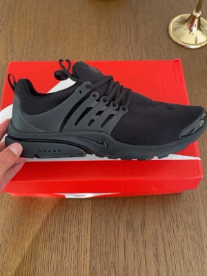 Svarta Nike Presto sneakers - Svarta Nike Presto sneakers med stilren design och elastisk ovandel i mesh. Skorna har en unik snörning och plastdetaljer på sidorna för extra stöd. Sulan är svart och ger bra komfort. Perfekta för dig som gillar en clean och sportig look.