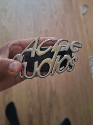 Acne Studios svart skärp i skinn - Snyggt svart skärp från Acne Studios med bred rem i skinn och en stor, silverfärgad buckle med Acne Studios-loggan i metallic. Perfekt statement piece för att lyfta vilken outfit som helst.