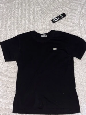 Svart t-shirt från Lacoste - Klassisk svart t-shirt från Lacoste med den ikoniska krokodilloggan broderad på bröstet. T-shirten har rund hals och korta ärmar, perfekt för en clean och stilren look. Tillverkad i mjuk bomull för maximal komfort.