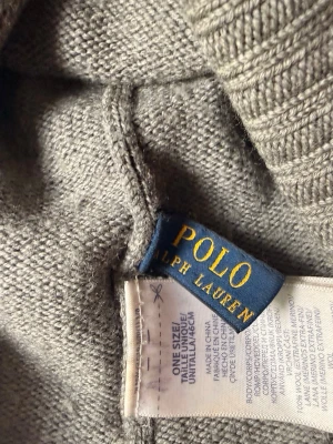 Grå stickad mössa Polo Ralph Lauren - Snygg grå stickad mössa från Polo Ralph Lauren. Tillverkad i mjukt material med klassisk ribbstickad kant. Perfekt accessoar för att hålla värmen och ge en stilren touch till din outfit.