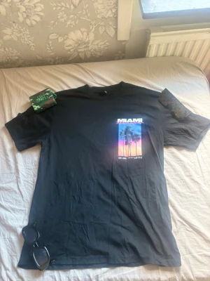 Svart Miami t-shirt med tryck - Säljer en svart t-shirt med färgstarkt Miami-tryck på bröstet. Motivet visar palmer mot en solnedgång i blått, rosa och orange. T-shirten har normal passform och är kortärmad, perfekt för dig som gillar streetstyle och grafiska prints.Pris kan diskuteras.