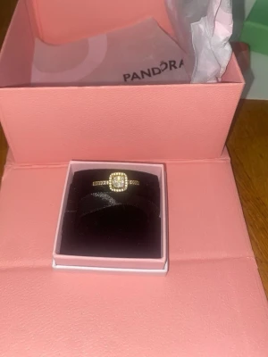 Guldfärgad ring med gnistrande stenar - Snygg guldfärgad ring från Pandora med en fyrkantig, gnistrande sten i mitten och små klara stenar runt om hela ringen. Perfekt för dig som gillar eleganta och trendiga accessoarer. Kommer i originalask.