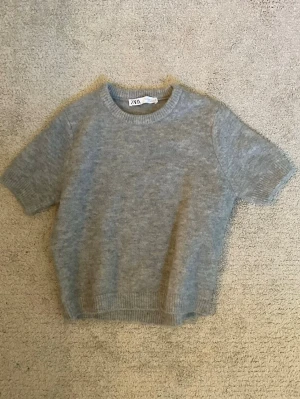 Grå stickad t-shirt  - Mysig grå stickad t-shirt från ZARA med korta ärmar och rund halsringning. Tröjan har ribbade muddar vid ärmslut och nederkant. Perfekt för lager på lager och enkel att styla till olika outfits.