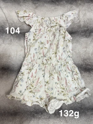 Blommig jumpsuit med volangärm - Supersöt vit jumpsuit med blommigt mönster i rosa, grönt och blått. Volangärmar och resår i midjan ger en skön passform. Perfekt för varma dagar och har fina spetsdetaljer vid bensluten.🌸 Fint använt skick. Storlek 104