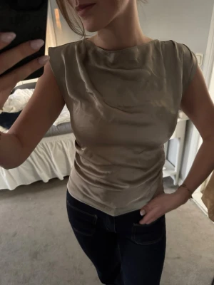Beige satinärmlös topp - Snygg beige topp i satin med draperad halsringning och ärmlös design. Toppen har en elegant, mjuk glans och smickrande passform. Perfekt att styla med jeans eller kjol för en trendig look.