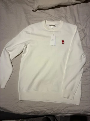 Vit stickad tröja från Ami Paris - Säljer en stilren vit stickad tröja från Ami Paris med rund halsringning och det ikoniska röda Ami de Coeur-emblemet broderat på bröstet. Tröjan har långa ärmar och ribbade muddar. Perfekt för dig som gillar minimalistisk design.