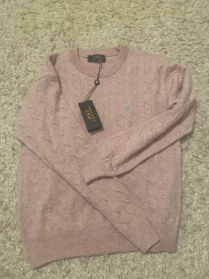 Ljusrosa stickad tröja Polo Ralph Lauren - Ljusrosa stickad tröja från Polo Ralph Lauren med klassiskt kabelstickat mönster och rund halsringning. Tröjan har lång ärm och den ikoniska broderade loggan på bröstet. Perfekt för en stilren och avslappnad look.