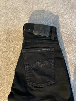 Svarta skinny jeans från Nudie Jeans - Säljer ett par svarta skinny jeans från Nudie Jeans med klassisk femficksdesign och diskret logga på bakfickan. Jeansen har en smal passform och är tillverkade i stretchigt material för extra komfort.