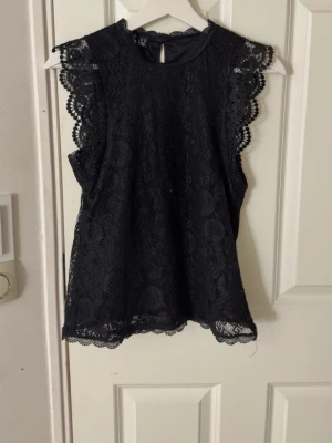 Svart spetsblus från Pieces XS - Supersnygg svart blus från Pieces i spets med blommigt mönster. Ärmlös modell med fina volangdetaljer längs axlarna och knapp i nacken. Perfekt att styla med jeans eller kjol för en trendig look.