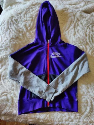 Lila Nike hoodie med grå ärmar/xl barn - Nike hoodie i lila med grå ärmar och svart magficka. Dragkedjan är röd och huvan är lila. Nike-logga i vitt på bröstet. Perfekt för en sportig och avslappnad stil.