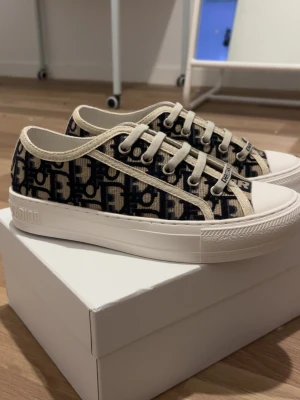 Dior sneakers - Snygga Dior Oblique sneakers i lågt snitt med beige och marinblått monogrammönster. Skorna har vita gummisulor, beige snörning och detaljer samt tydlig Dior-logga på sidan och hälen. Perfekta för dig som vill ha en lyxig och trendig look.