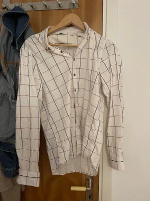 Vit rutig henley-skjorta - Vit henley-skjorta med rutigt mönster i mörkblått och brunt. Skjortan har lång ärm, knappar framtill och klassisk krage. Perfekt för en avslappnad men ändå stilren look.