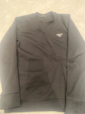 Svart sweatshirt från Prada - Svart sweatshirt från Prada med rund halsringning och lång ärm. Tröjan har en ikonisk metallogo på bröstet och är tillverkad i mjukt material. Enkel och stilren design som ger en exklusiv känsla.