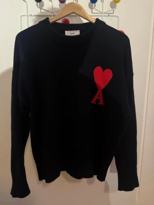 Svart stickad tröja från Ami - Svart stickad tröja från Ami Alexandre Mattiussi med röd hjärt- och A-detalj på bröstet. Tröjan har rund halsringning och långa ärmar. Perfekt för dig som gillar stilrena plagg med en twist.