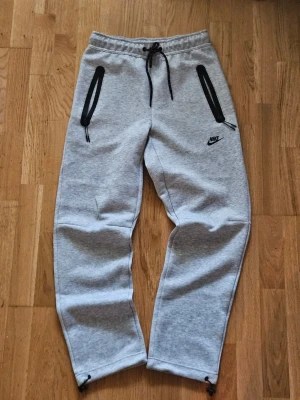 Grå baggy Nike tech byxor - Snygga grå mjukisbyxor från Nike med svarta dragkedjor på fickorna och justerbar dragsko i midjan. Byxorna har en avslappnad passform och är tillverkade i mjukt material. Nike-logga broderad på vänster ben. Perfekta för chill eller träning.