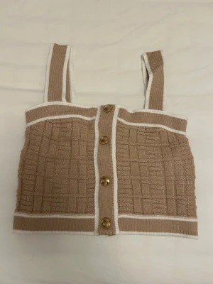Beige stickad crop top med knappar från SHEIN - Beige stickad crop top med breda axelband och vita kanter från SHEIN i storlek XS. Toppen har ett rutigt mönster och fyra dekorativa knappar framtill. 