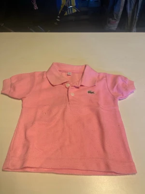 Rosa pikétröja från Lacoste - Snygg rosa pikétröja från Lacoste med klassisk krage och två knappar framtill. Tröjan har korta ärmar och den ikoniska krokodilloggan broderad på bröstet. Perfekt för en avslappnad och sportig look.