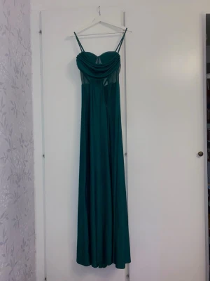 Faeriesty bal-och långklänning  - Elegant mörkgrön långklänning med smala dubbla axelband och draperad byst. Klänningen har en figurnära överdel och ett flytande, utsvängt kjolparti. Perfekt för speciella tillfällen och har en modern, stilren look.