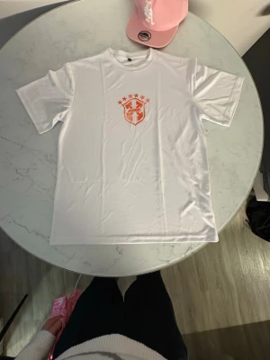 Vit t-shirt från Asedunk med tryck - Vit t-shirt från Asedunk med orange tryck framtill i form av ett sköldmärke och stjärnor. Klassisk passform med rund hals och korta ärmar. Perfekt för en sportig och avslappnad stil.