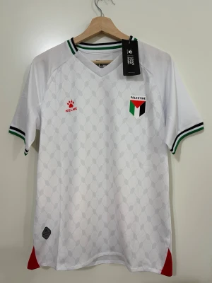 Vit Palestina fotbollströja Kelme - Snygg vit fotbollströja från Kelme med Palestina-emblem på bröstet och diskret mönster över hela tröjan. Ribbade muddar och krage med gröna, svarta och röda detaljer. Perfekt för dig som vill visa stöd på matchen eller träningen.