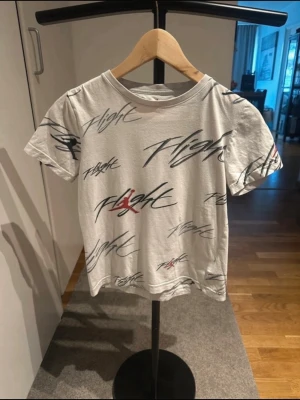 Vit Jordan Flight t-shirt 10-12år - Säljer en vit t-shirt från Jordan med svart och röd 'Flight'-text tryckt över hela tröjan. Klassisk rund hals och korta ärmar, perfekt för en sportig och avslappnad stil.